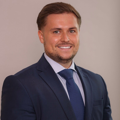 A man smiles while wearing a dark suit, light blue shirt, and blue tie; standing against a plain light-colored background.