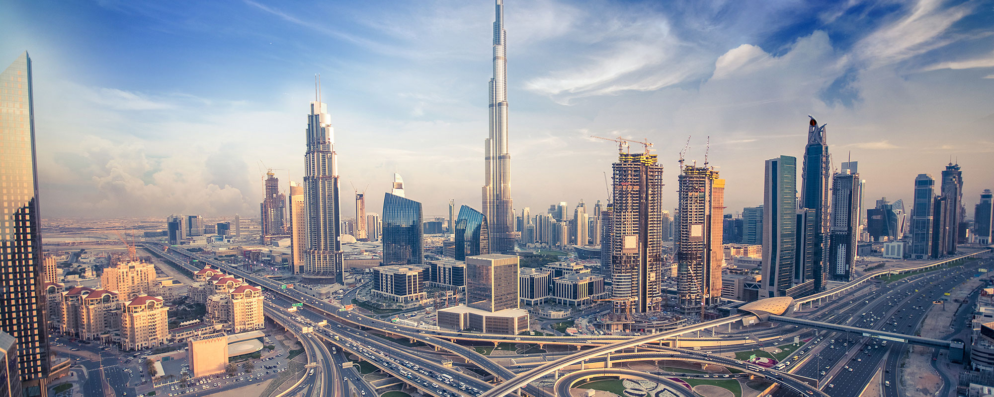Skyscrapers rise above busy highways in a sprawling urban landscape during the day, with partial cloud cover and construction cranes visible in the background.