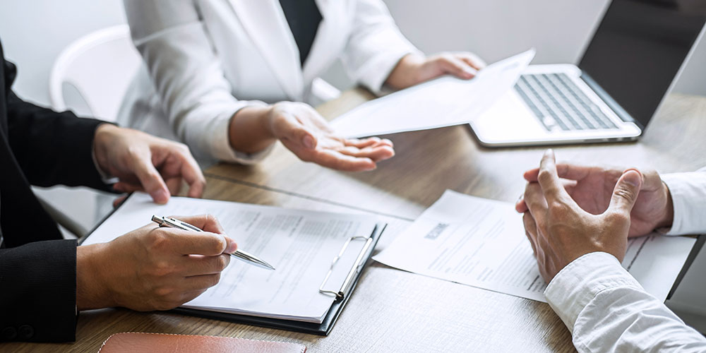 Three individuals in business attire engage in a discussion, gesturing over documents. A laptop rests nearby on a wooden desk, suggesting a professional meeting setting.