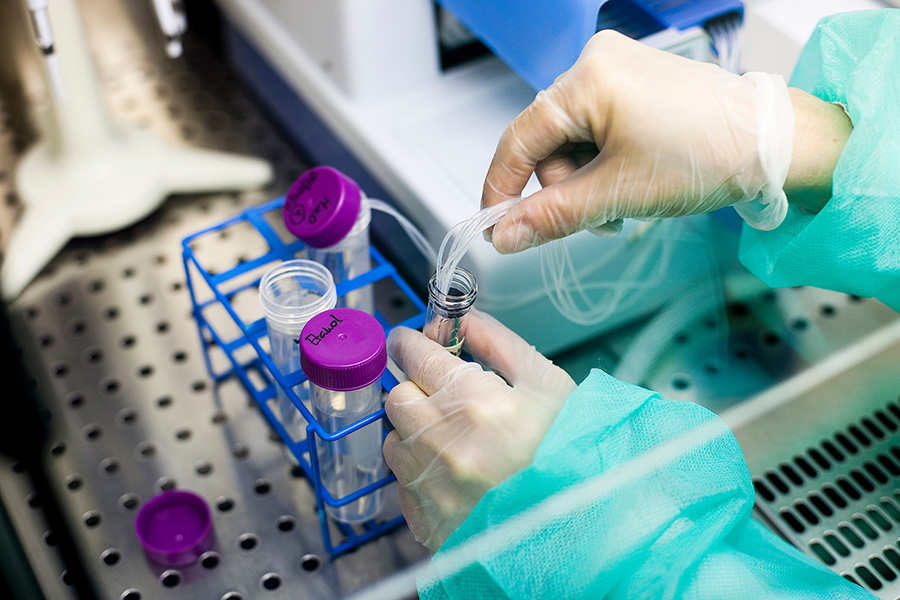 Gloved hands manipulate a tube, transferring liquid via a thin pipette. Nearby, a rack holds labeled test tubes with purple caps, labeled 