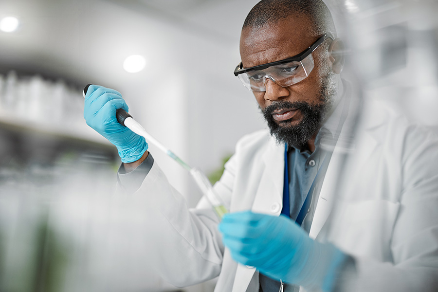A scientist wearing safety goggles and blue gloves uses a pipette to transfer liquid into a test tube in a laboratory setting.