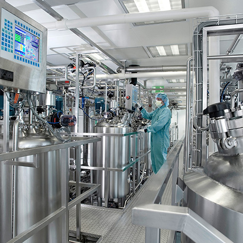 A person wearing protective clothing operates machinery in an industrial facility, surrounded by large stainless steel tanks and interconnected pipes. A control panel displays various settings and data.