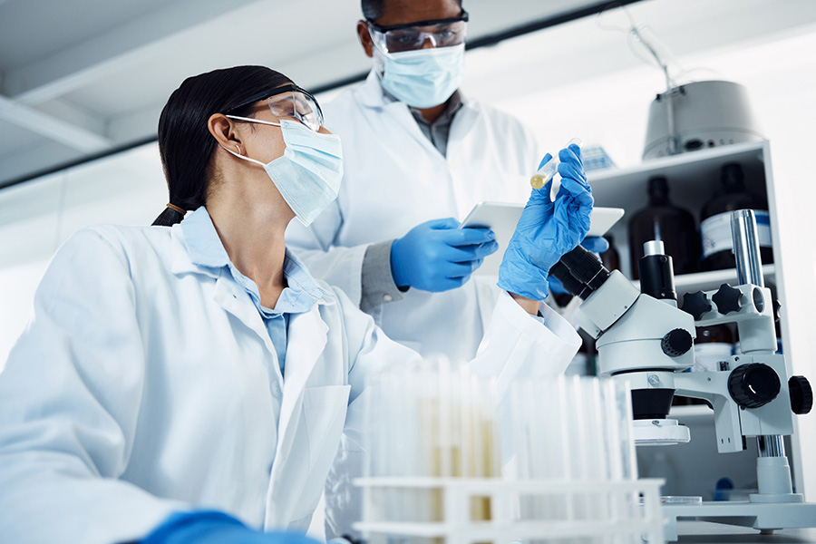 Scientists work collaboratively in a laboratory, examining a test tube by a microscope. They wear lab coats, gloves, masks, and goggles, surrounded by shelves with bottles and laboratory equipment.