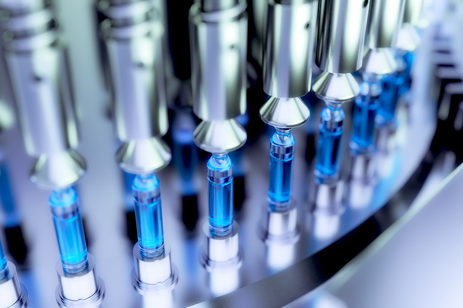 Glass vials, filled with blue liquid, are arranged on a rotating industrial platform, prepared for filling or capping in a pharmaceutical manufacturing setting.