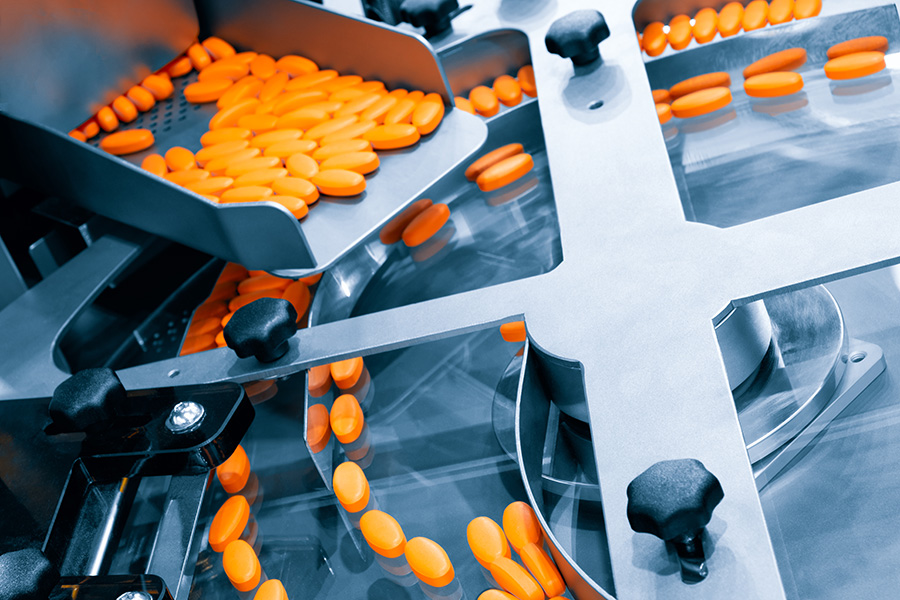 Orange pills are moving through a metal pill-sorting machine on a conveyor belt, set within a pharmaceutical manufacturing facility.