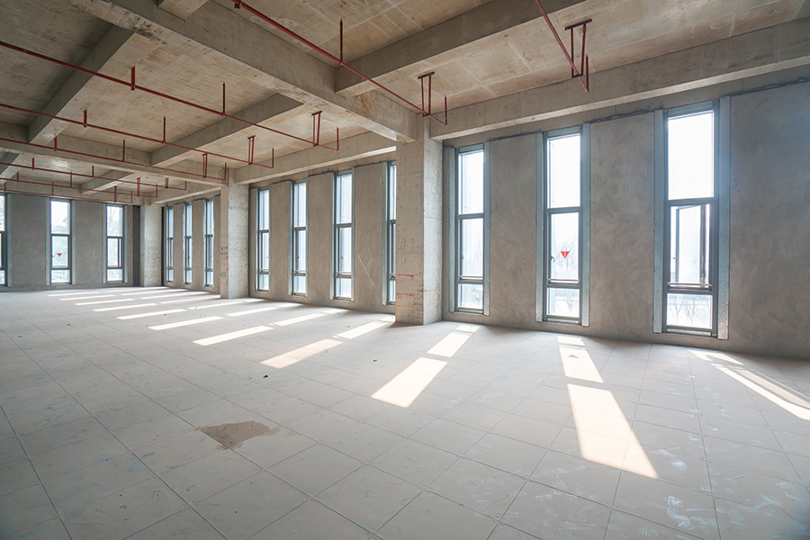 An empty, unfinished room with large windows admits sunlight, casting shadows across the tiled floor. Exposed red pipes run along the ceiling in a minimal, industrial setting.