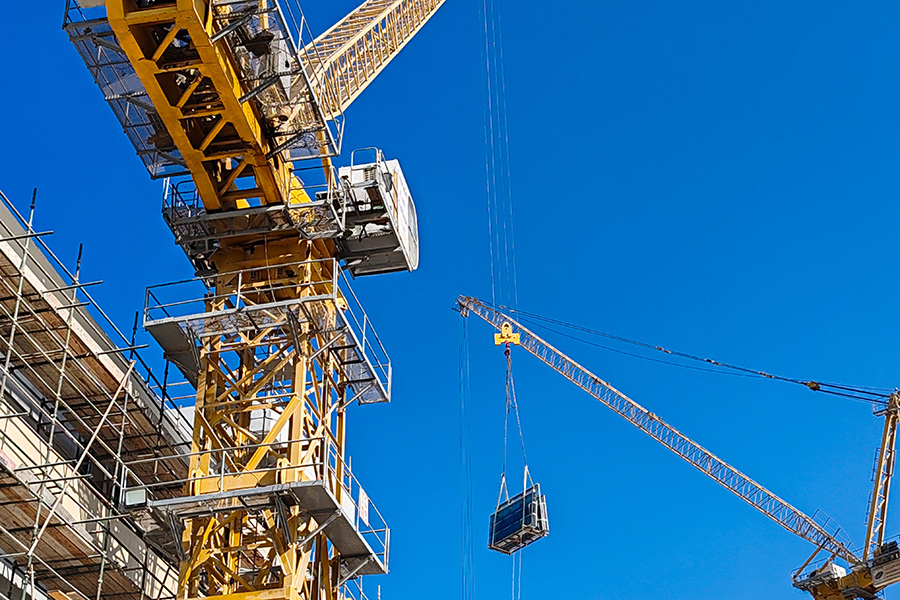 A yellow construction crane lifts a metal structure beside a building under scaffolding, framed against a clear blue sky.