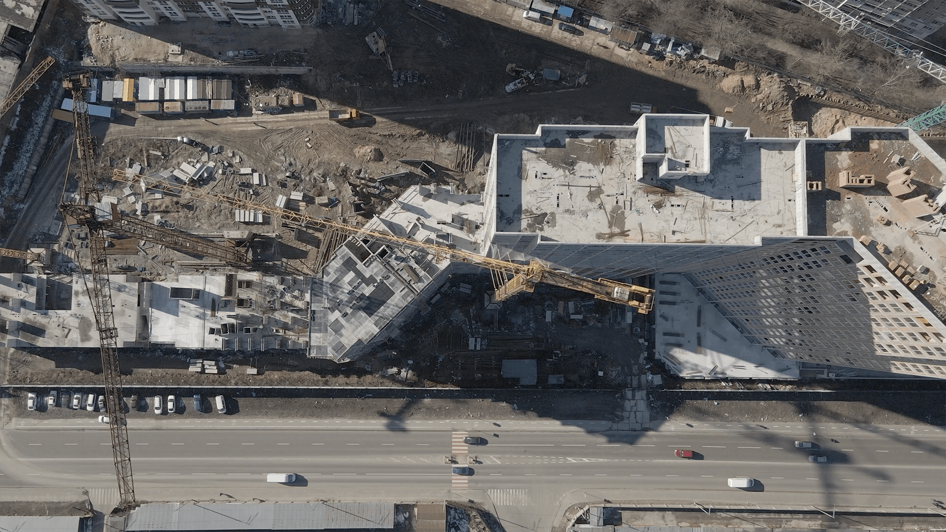 A construction crane positions materials on a partially built structure, surrounded by dirt and equipment, beside a road with moving cars.