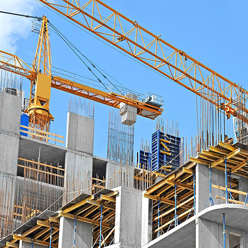 A yellow crane lifts materials at a construction site, surrounded by partially built concrete structures with scattered metal rods, under a clear blue sky.