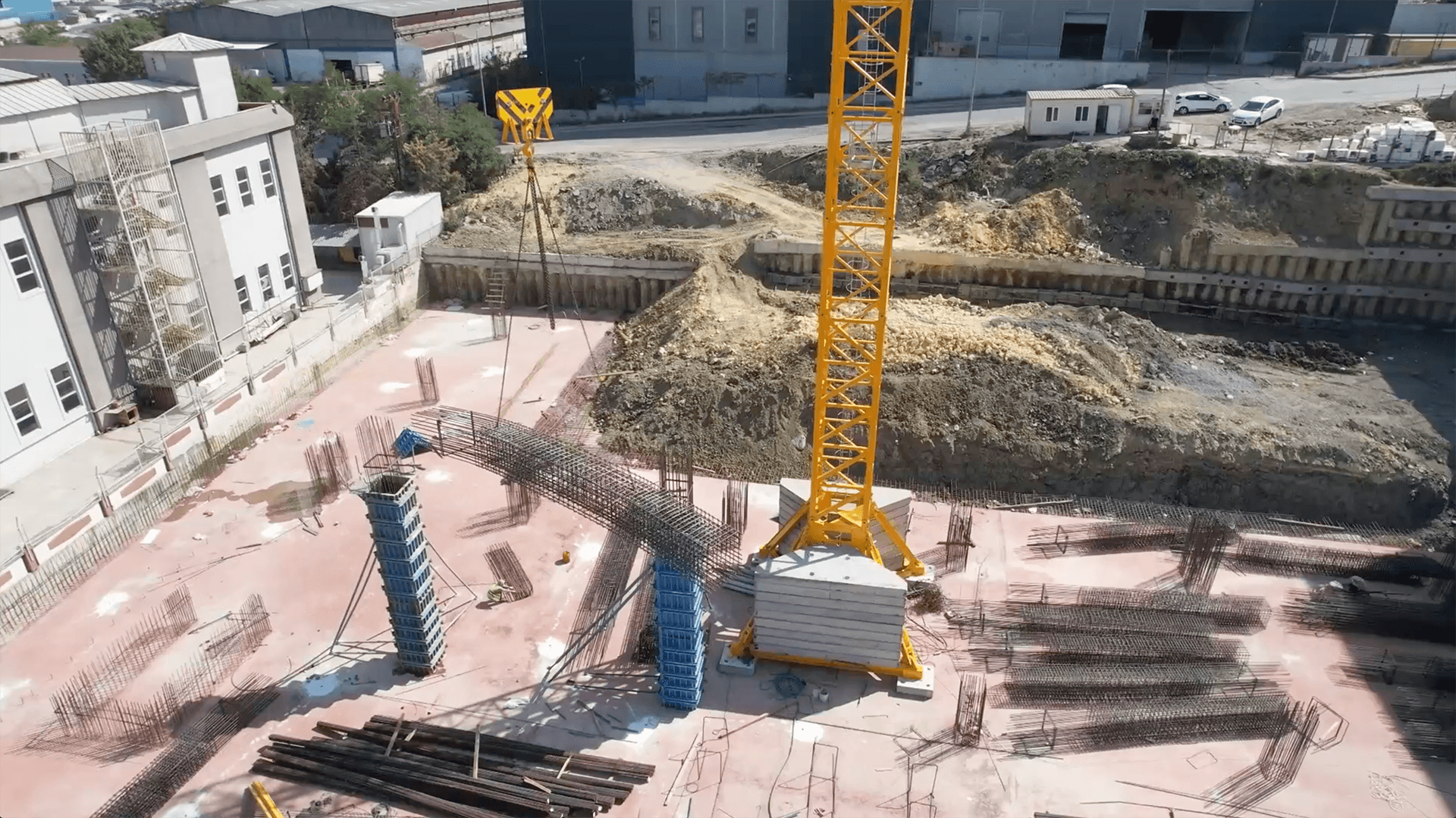 construction site with large cranes building a multi-story structure. The site is surrounded by equipment and materials, adjacent to a busy road with moving vehicles.