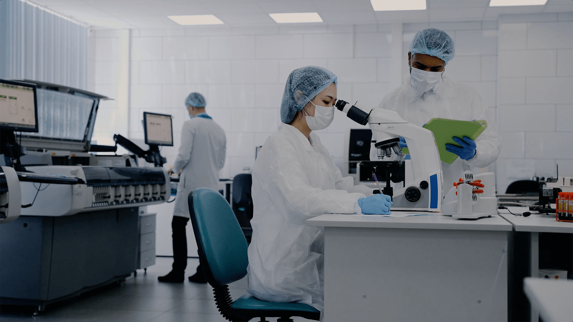Scientists, wearing protective gear, work in a laboratory. One looks through a microscope, while another reviews a tablet. The lab is equipped with computers and testing equipment.