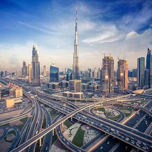 Skyscraper towers above a sprawling urban landscape, surrounded by interconnected highways filled with moving vehicles, while modern buildings stand tall under a bright, partly cloudy sky.