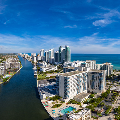 Skyscrapers line a waterfront, bordered by a canal on one side and ocean on the other, under a clear blue sky. Nearby, a swimming pool and low-rise buildings complete the scene.