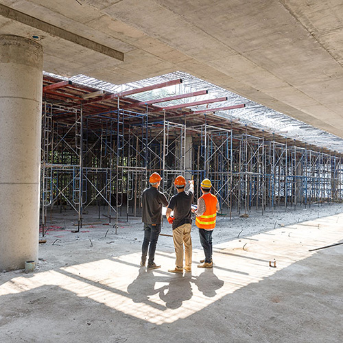 Three construction workers, wearing helmets and vests, stand discussing under a partially constructed concrete ceiling, surrounded by scaffolding and sunlight.