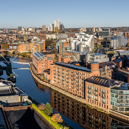 Red-brick buildings line a riverbank, reflected in the water. Urban skyline features modern and historic architecture under a clear blue sky, suggesting a bustling cityscape.