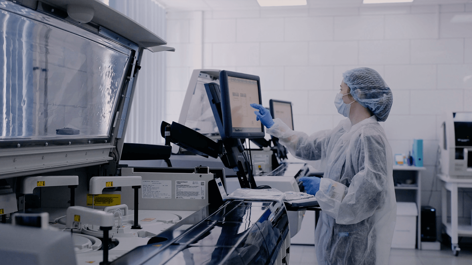A lab technician in protective gear uses a touch screen monitor, surrounded by machinery in a sterile laboratory environment.