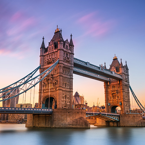 A historic bridge towers over a calm river, with gothic-style architecture and suspension elements glowing in warm evening light, against a backdrop of a colorful sky and urban skyline.