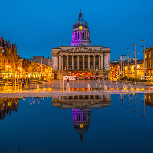 A grand building with a domed roof and illuminated columns reflects in a tranquil water feature. The plaza is surrounded by vibrant city lights and evening sky.