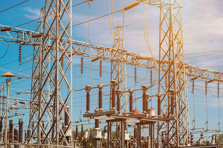 Electrical substation structures stand interconnected, supporting insulators and power lines against a clear blue sky with sunlight casting a warm glow over the metal framework.