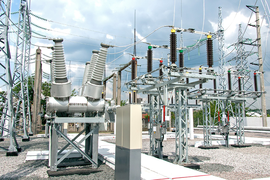 High-voltage electrical equipment stands on gravel, surrounded by metal structures and wires, in an open-air power substation under a cloudy sky.