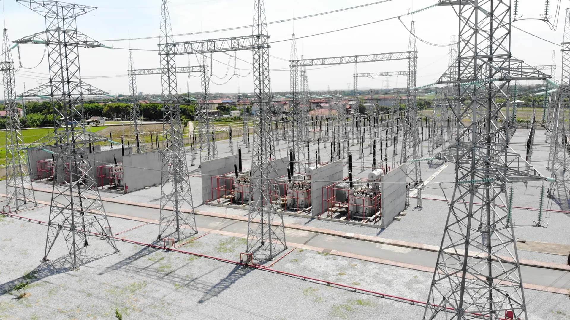 Steel transmission towers support wires, interconnecting electrical transformers in an expansive industrial substation surrounded by fields and distant buildings under a clear sky.