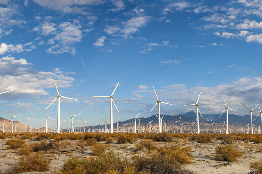 Wind turbines rotate in a desert landscape, surrounded by dry bushes under a bright, cloudy sky. Mountains rise in the background, creating a vast and open setting.