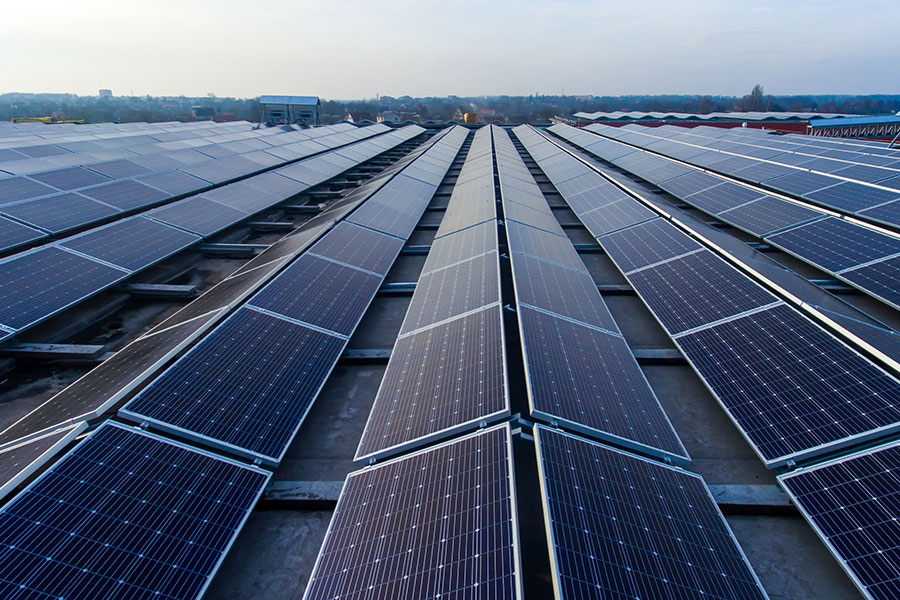 Rows of solar panels are installed on a flat rooftop, capturing sunlight against a partly cloudy sky, with a distant treeline on the horizon.