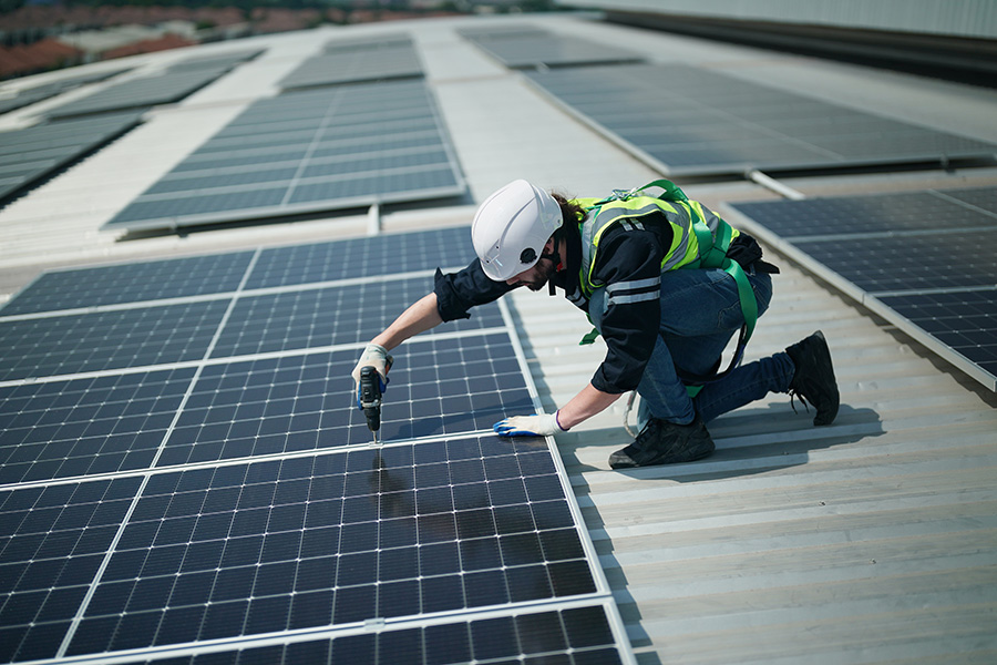 A worker in safety gear is installing solar panels on a large rooftop, using a power drill amidst a series of aligned panels under bright daylight.