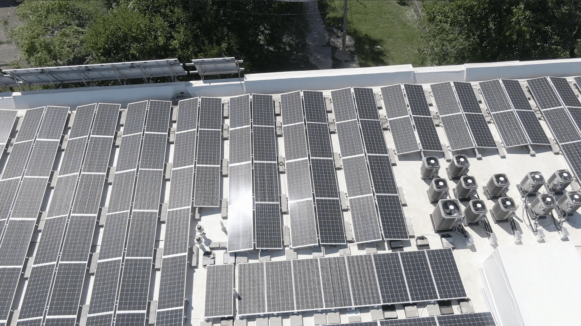 Solar panels cover a rooftop, capturing sunlight, while multiple air conditioning units are clustered nearby; surrounded by trees, suggesting an eco-friendly urban setting.